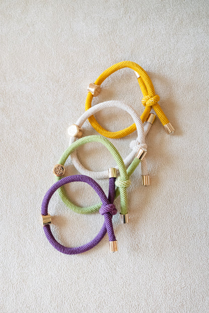 Four hair ties in purple, green, white, and yellow with gold accents, stacked in a cascade arrangement on a beige surface, showcasing their knotted design.