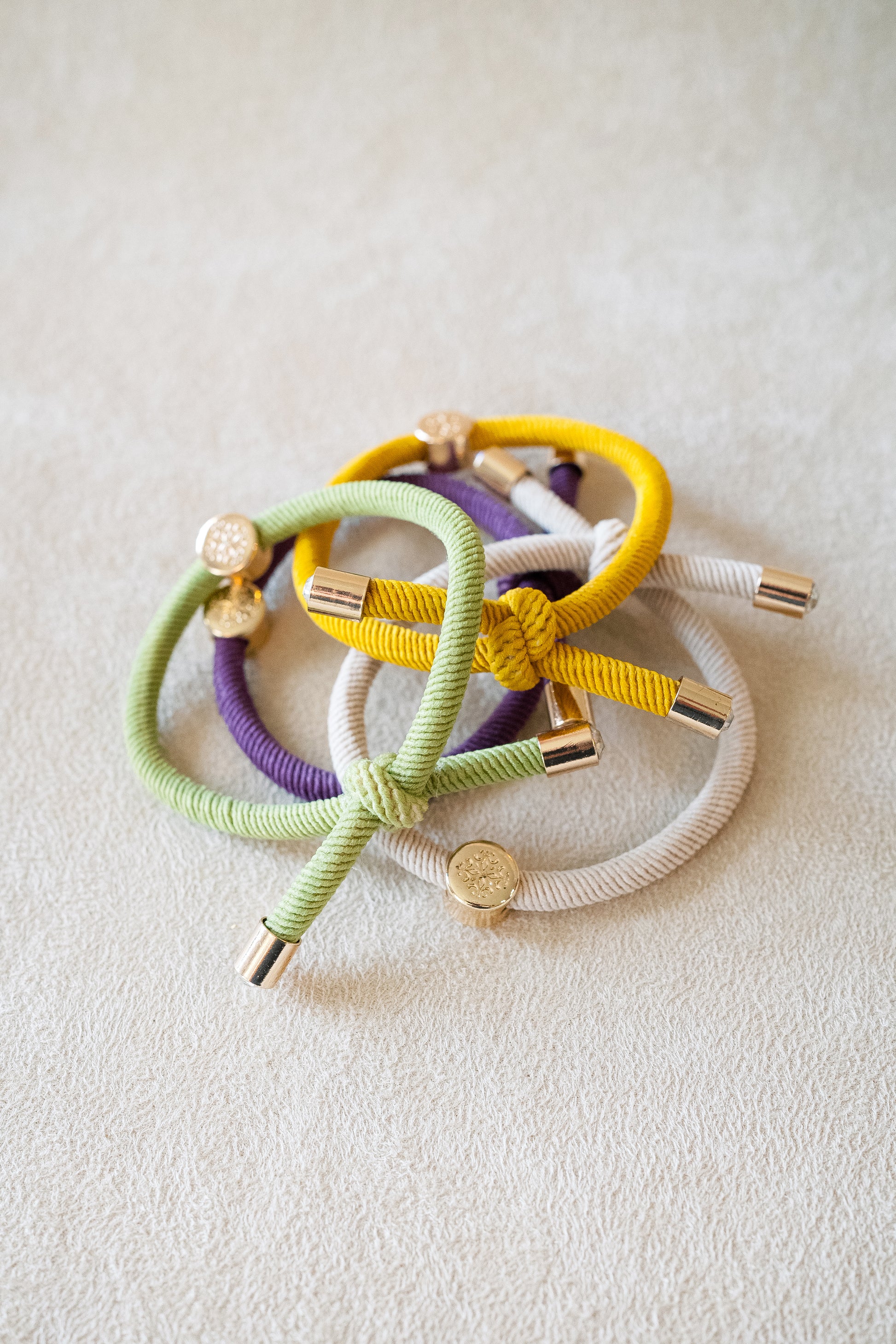 Four elastic hair ties in green, yellow, purple, and beige with gold accents, casually arranged in a cluster on a beige surface, highlighting their texture.