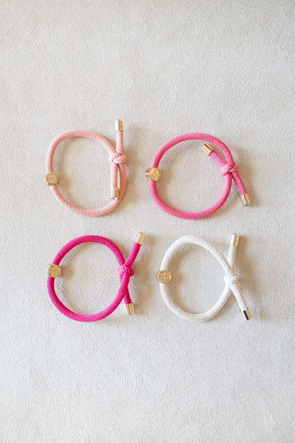 Four elastic hair ties in soft pink, vibrant pink, and white, arranged neatly in a grid on a light beige surface, each accented with shiny gold details.