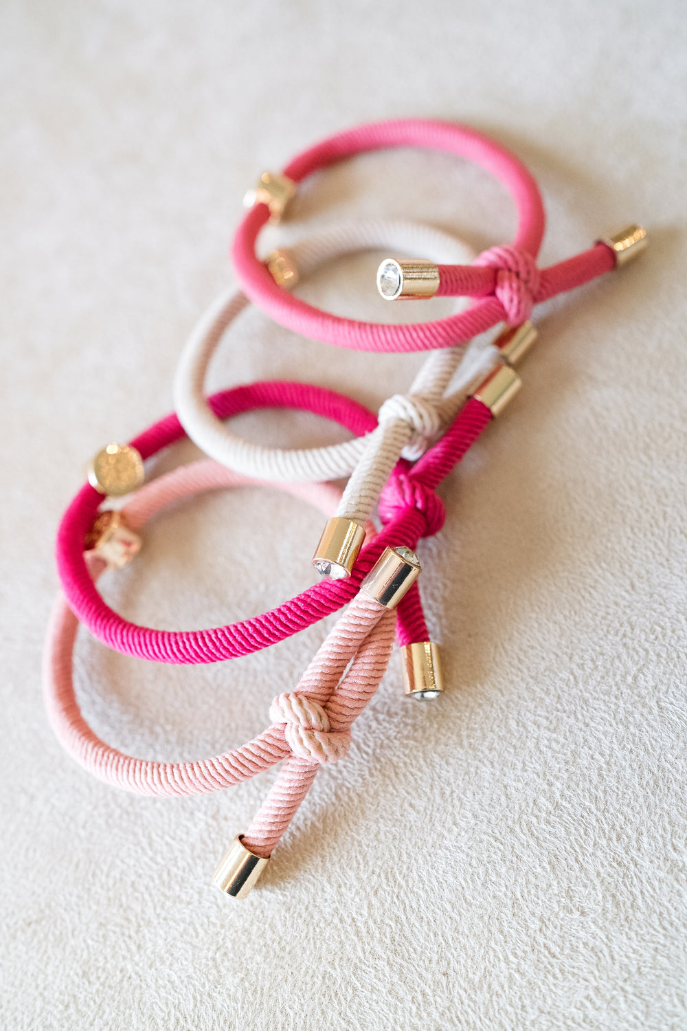 Four hair ties in shades of pink and beige with gold accents, arranged in a stack on a beige surface, showcasing their vibrant colors and metallic details.