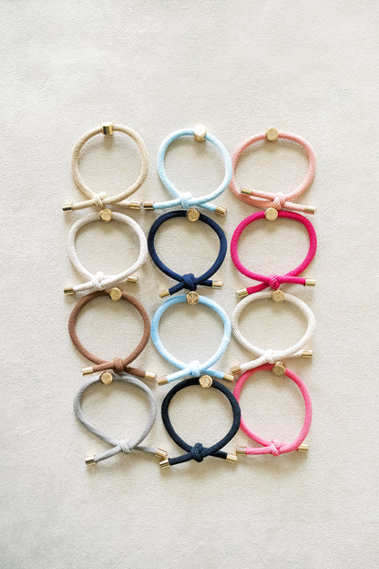 Twelve colorful elastic hair ties arranged in a neat grid pattern on a beige surface, featuring shades of blue, pink, brown, and beige with gold accents.