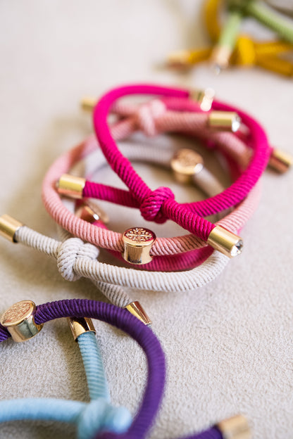 Close-up of stacked elastic hair ties in vibrant pink, soft beige, and purple, arranged on a light surface, with gold accents highlighting their texture.