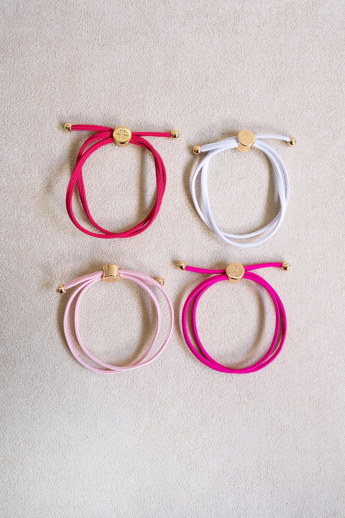 Four colorful, double-wrapped elastic hair ties, including red, white, light pink, and bright pink, arranged on a neutral background with gold accents