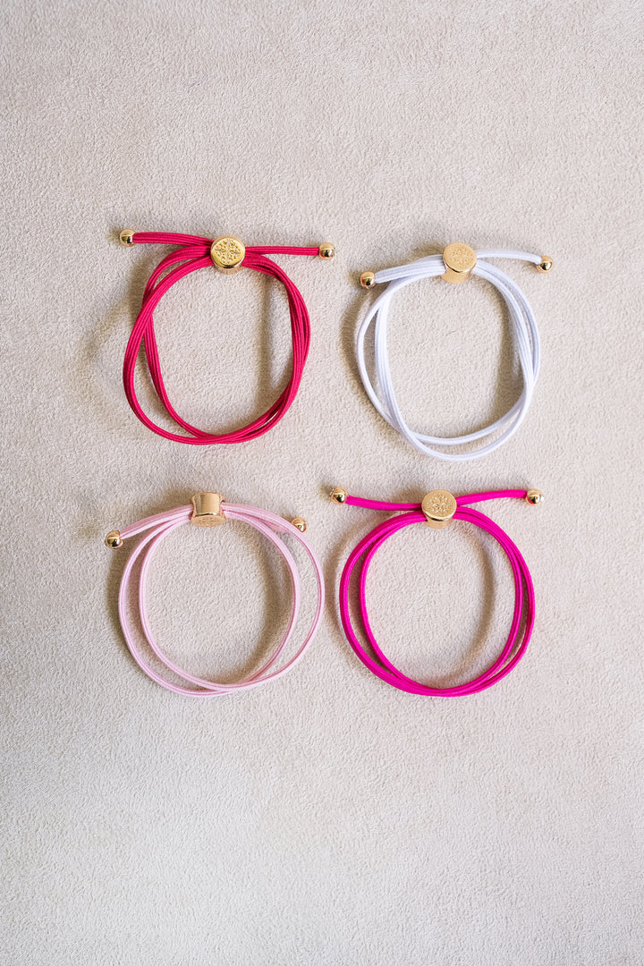 Four colorful, double-wrapped elastic hair ties, including red, white, light pink, and bright pink, arranged on a neutral background with gold accents