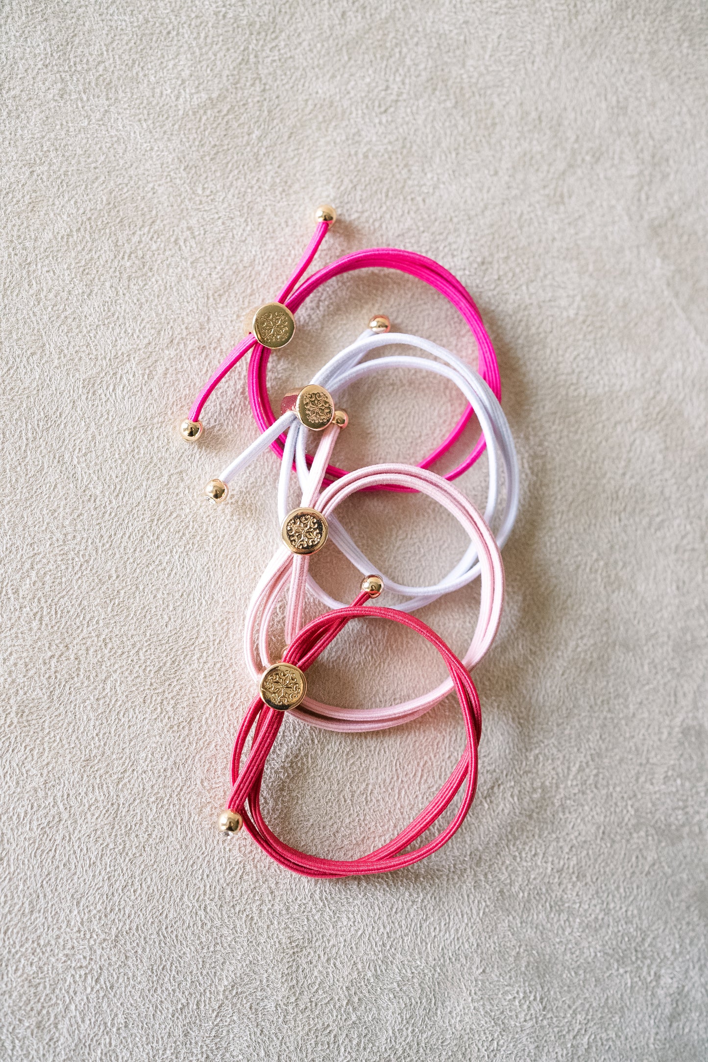 Four thin, stretchable hair ties are placed on a beige surface in red, white, pink, and fuchsia shades, accented with gold detailing.