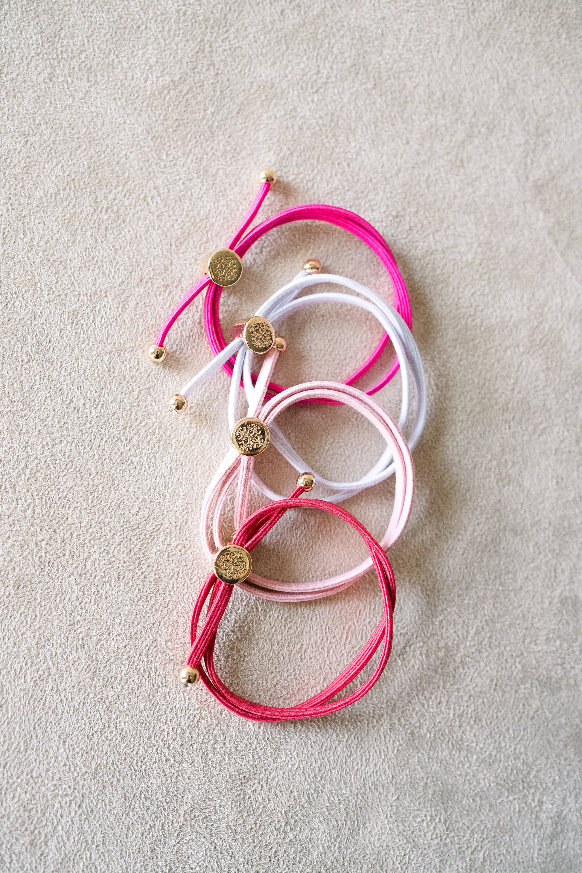 Four thin, stretchable hair ties are placed on a beige surface in red, white, pink, and fuchsia shades, accented with gold detailing.