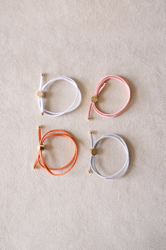 Four pastel-colored hair ties with gold accents are laid neatly on a beige surface, featuring colors like white, pink, orange, and gray arranged in a square.