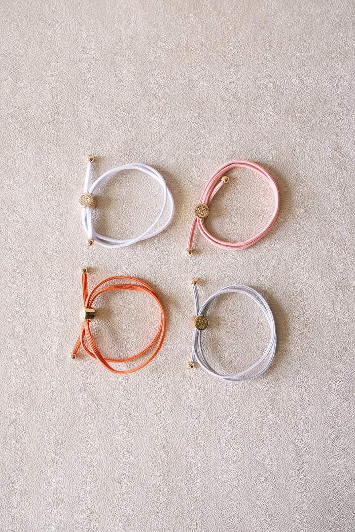 Four pastel-colored hair ties with gold accents are laid neatly on a beige surface, featuring colors like white, pink, orange, and gray arranged in a square.