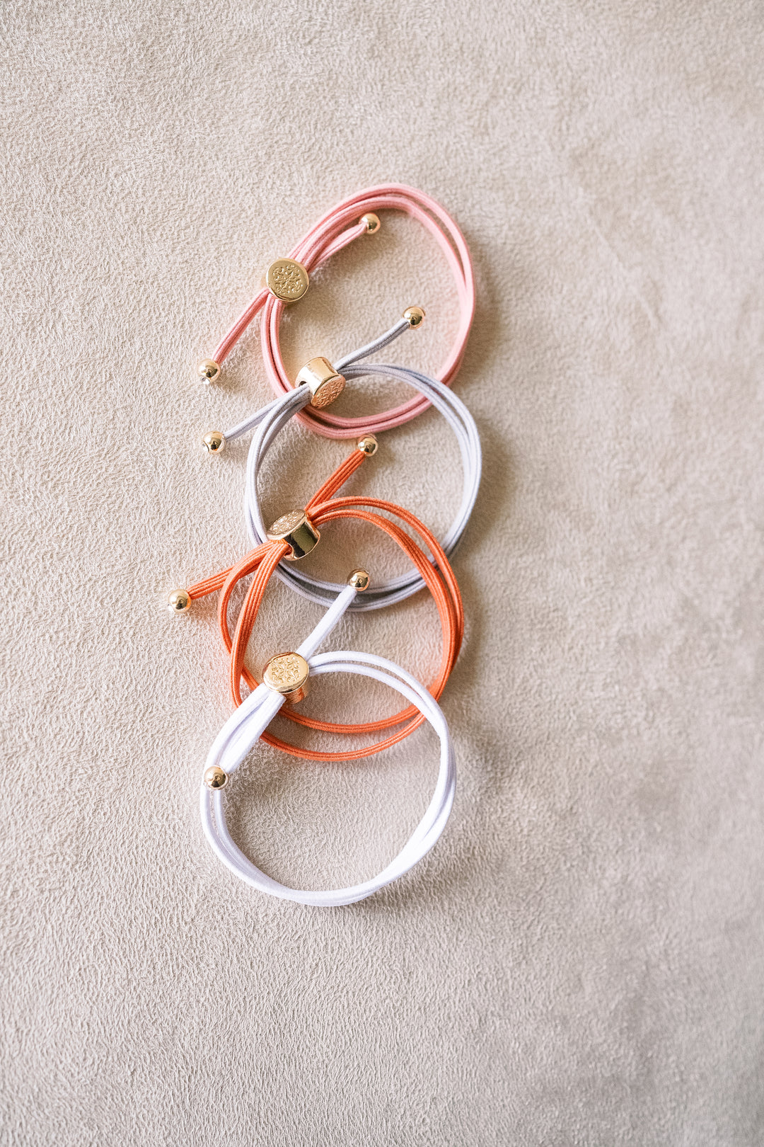 Four hair ties in peach, orange, grey and white with gold accents, stacked in a cascade arrangement on a beige surface, showcasing their wrapped design.