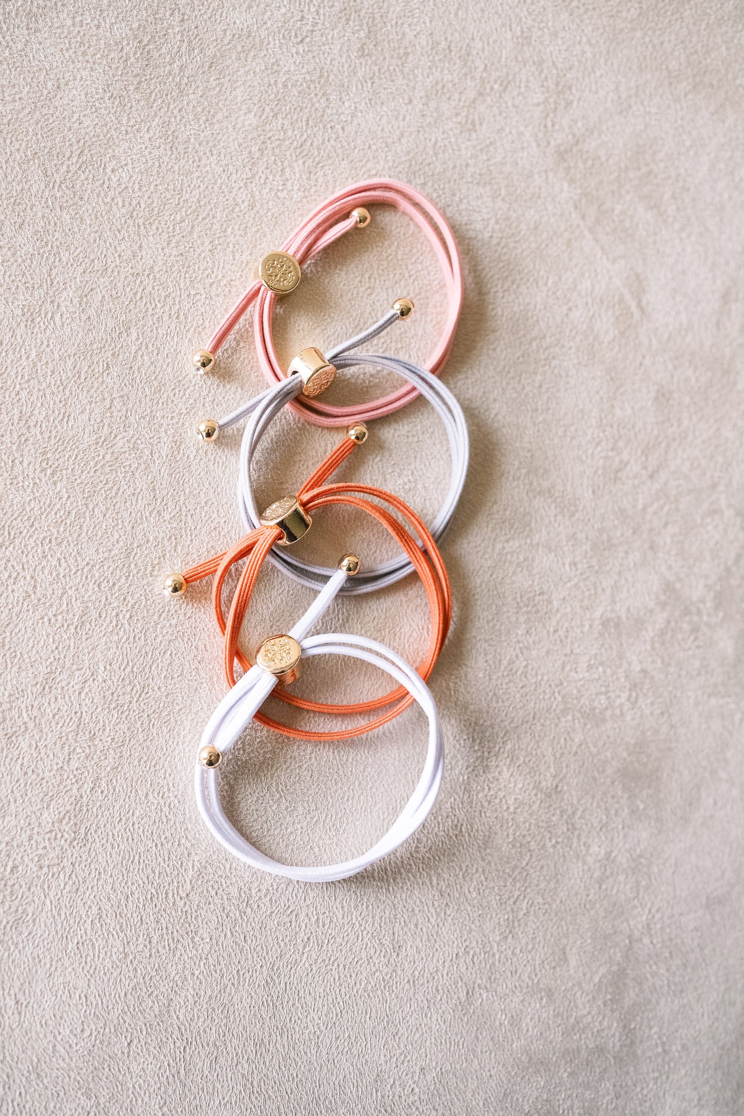 Four hair ties in peach, orange, grey and white with gold accents, stacked in a cascade arrangement on a beige surface, showcasing their wrapped design.