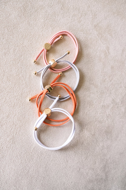 Four hair ties in peach, orange, grey and white with gold accents, stacked in a cascade arrangement on a beige surface, showcasing their wrapped design.