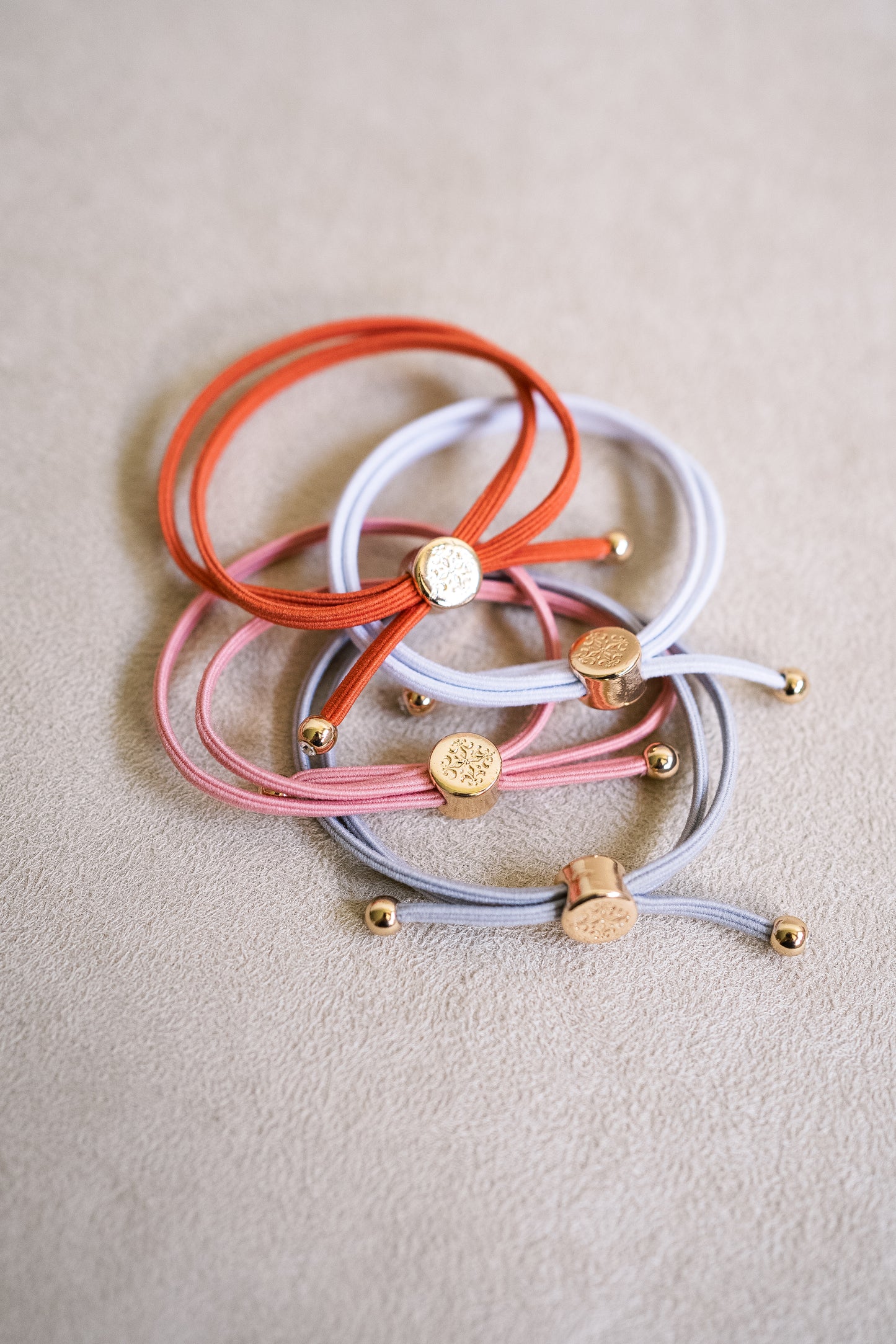 Four hair ties laid on a beige background, featuring colors like orange, pink, grey, and white, all adorned with gold-tone accents.