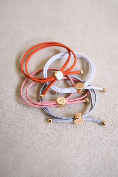 Four hair ties laid on a beige background, featuring colors like orange, pink, grey, and white, all adorned with gold-tone accents.