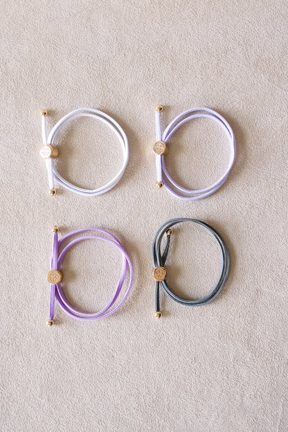 Four hair ties in pastel shades of white, lilac, lavender, and grey are arranged in a square pattern on a neutral, textured surface.