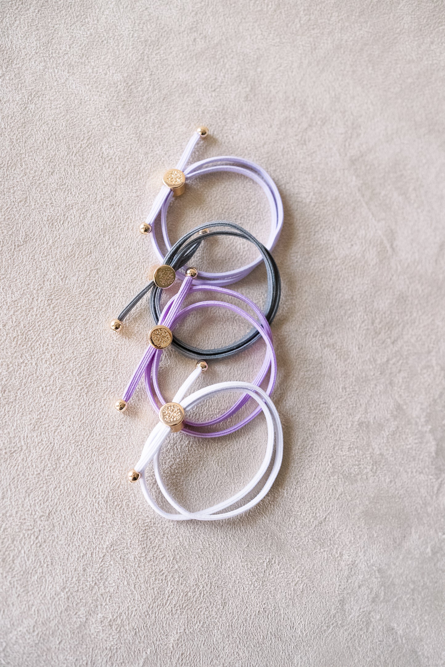 Four elastic bracelets in white, purple, and gray with round gold clasp and small gold beads at the ends are neatly arranged on a soft beige surface. 