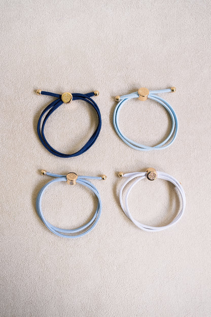 Four elastic hair ties in navy, various shades of blue, and white with gold accents arranged in a grid on a soft beige background.