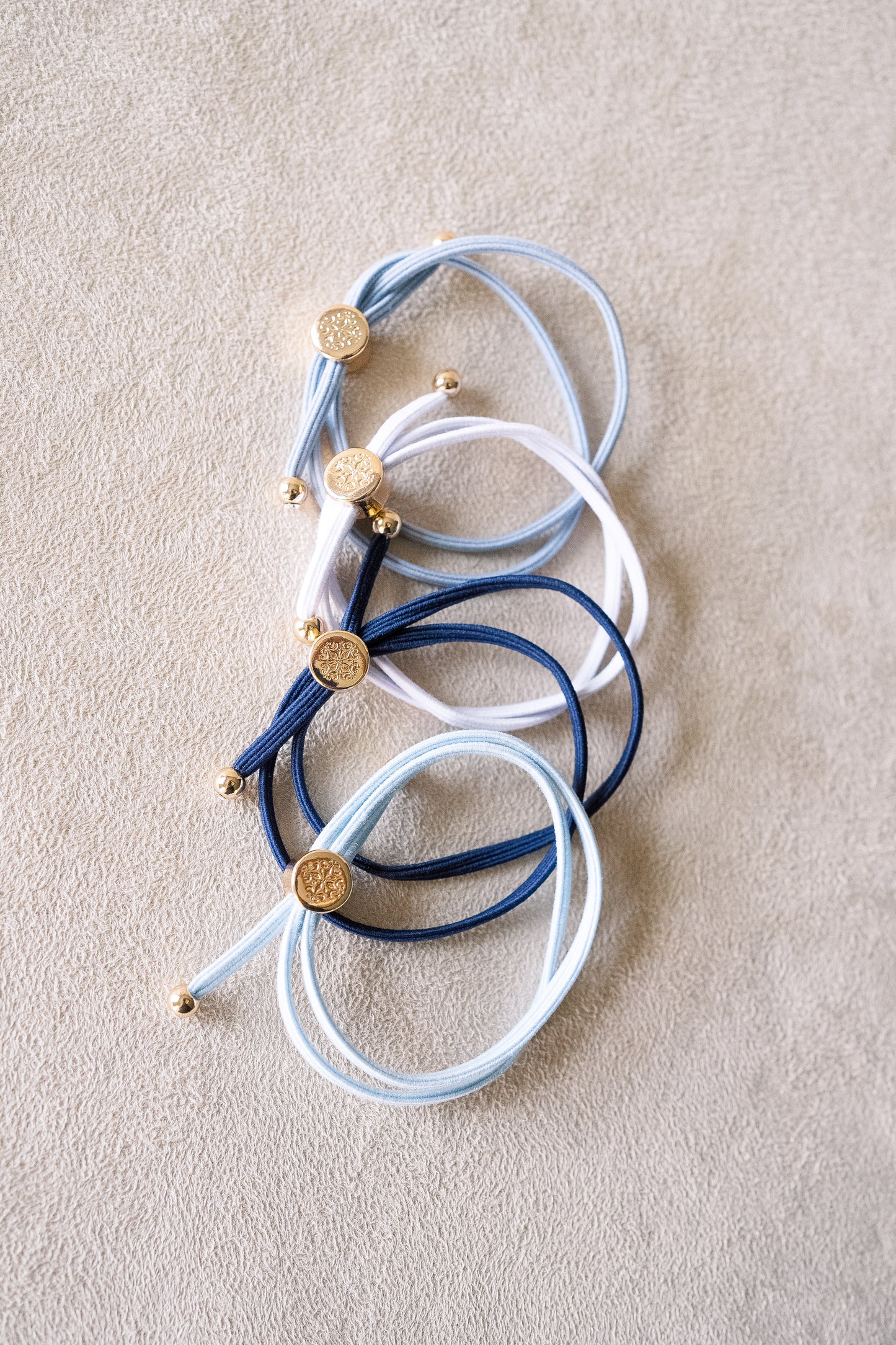 Four hair ties in navy, blue, light blue, and white with gold accents, stacked in a cascade arrangement on a beige surface, showcasing their wrapped design.