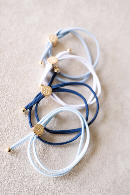 Close up shot of four hair ties in various shades of blue and white with gold accents, stacked in a cascade arrangement on a beige surface.