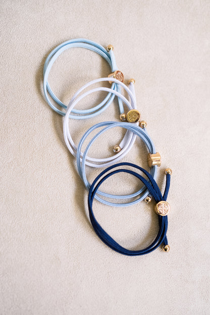 Four elastic double-wrapped hair ties in shades of navy, blue, and white with gold accents, neatly arranged on a textured beige surface.