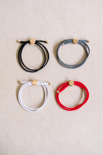 Four elastic hair ties in shades of bright red, white, grey, and black with gold accents arranged in a grid on a soft beige background.