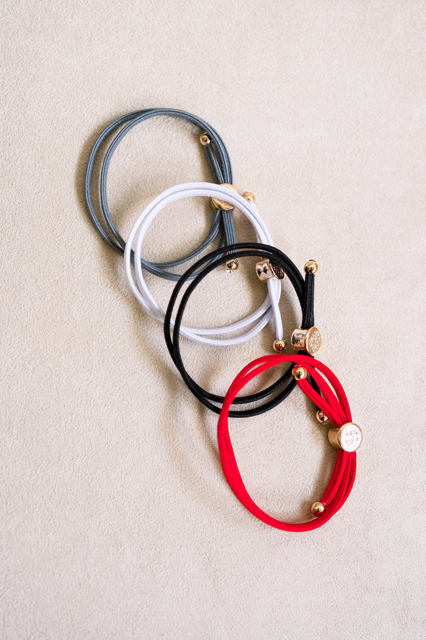 Four double-wrapped elastic hair ties in shades of gray, white, black, and red, featuring gold-toned accents, arranged on a neutral fabric background.