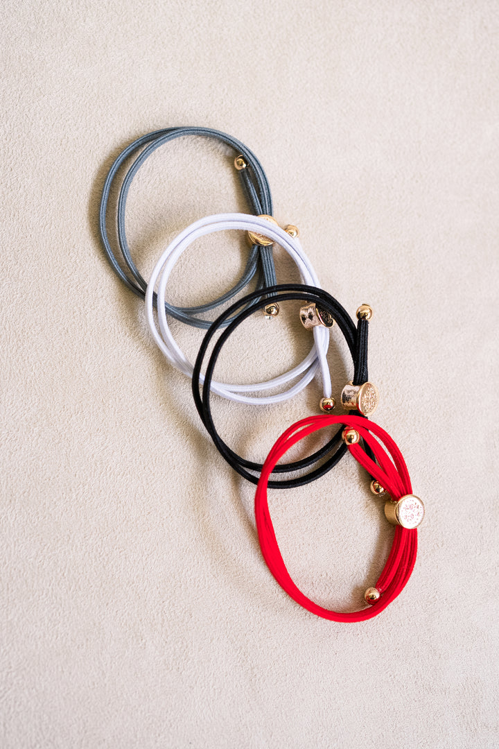 Four double-wrapped elastic hair ties in shades of gray, white, black, and red, featuring gold-toned accents, arranged on a neutral fabric background.