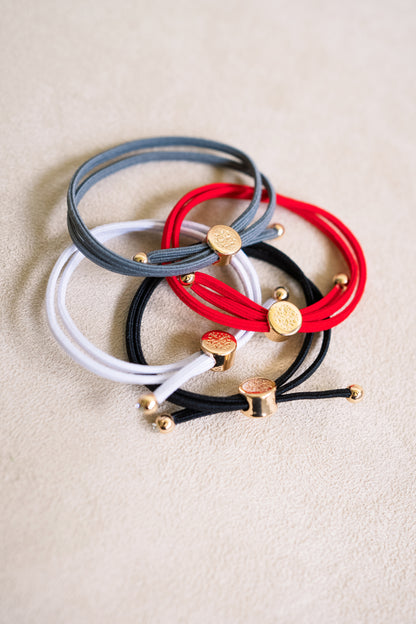 A close-up shot of four elastic bracelets in various colors—gray, red, black, and white—laid together on a beige surface, with gold embellishments.