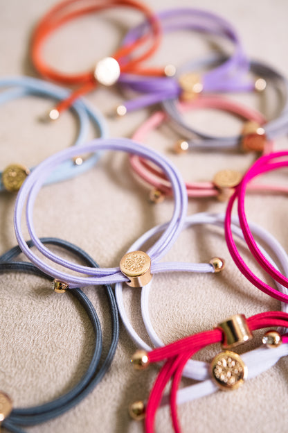 Colorful hair ties with a gold charm are arranged in a visually appealing layout. The ties vary in shades of lavender, orange, pink, and grey