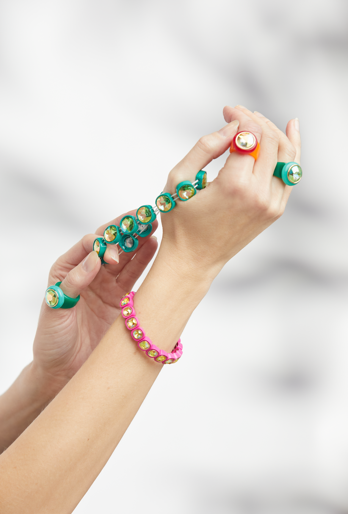 Two hands stretching a bright green bracelet with faceted gems, paired with a pink bracelet of similar design, and three orange and green oversized rings.