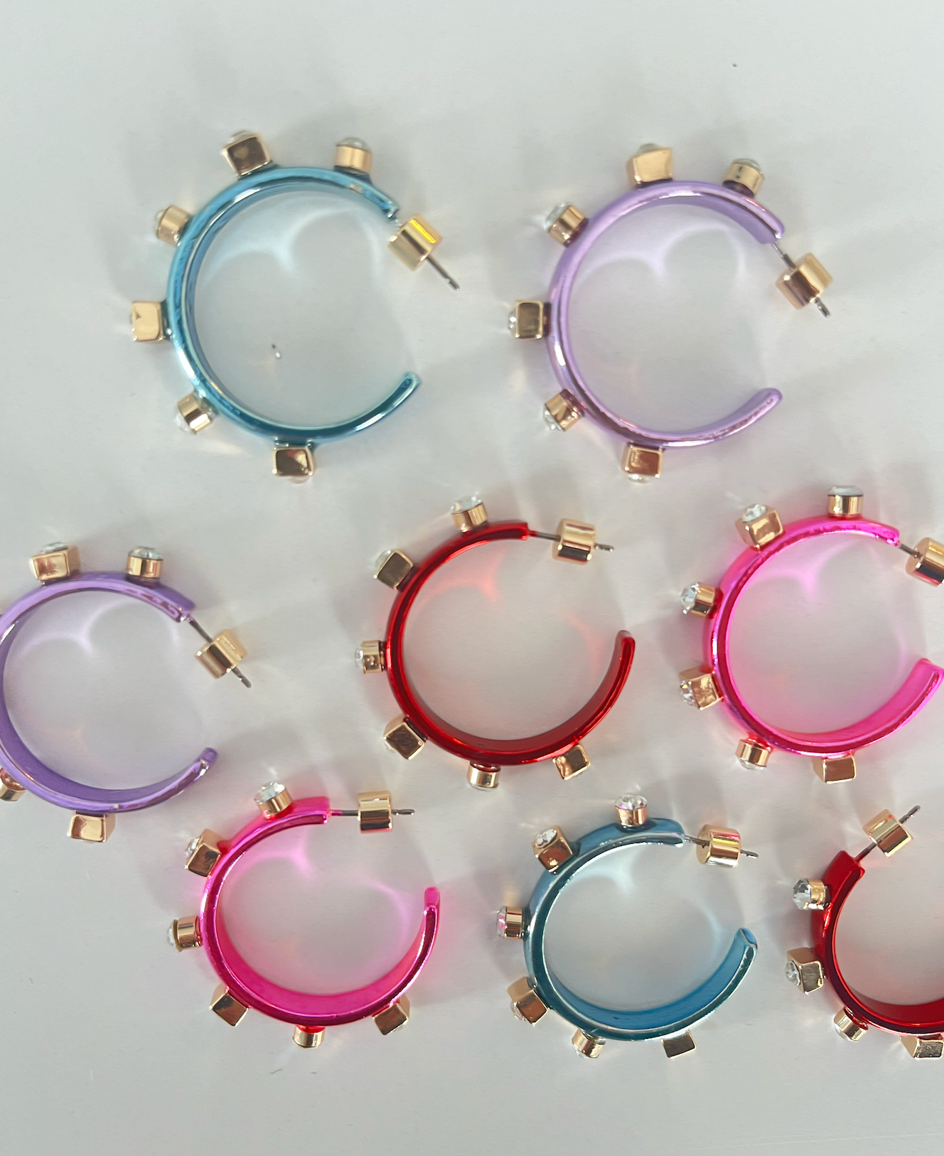 Colorful hoop earrings in blue, red, pink, and purple, adorned with gold studs and jewels, laid out on a flat surface with soft reflections.