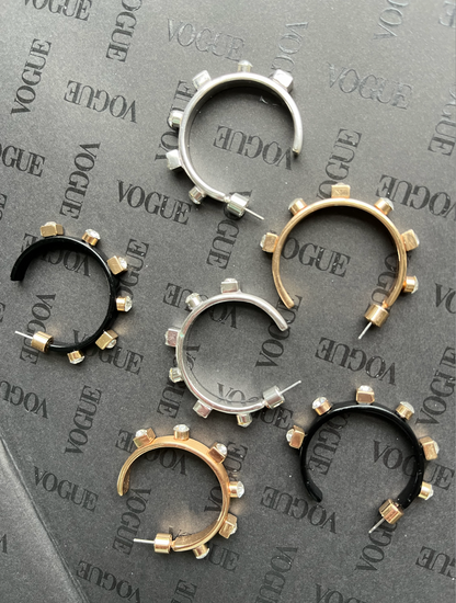 Hoop earrings in silver, gold, and black with metallic studs are arranged on a gray "Vogue" background, creating a sleek and stylish display.