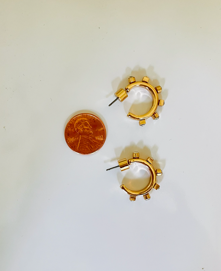 A pair of small, circular gold hoops with small, raised studs evenly spaced around their outer edges, placed next to a 2022 penny for size comparison. 