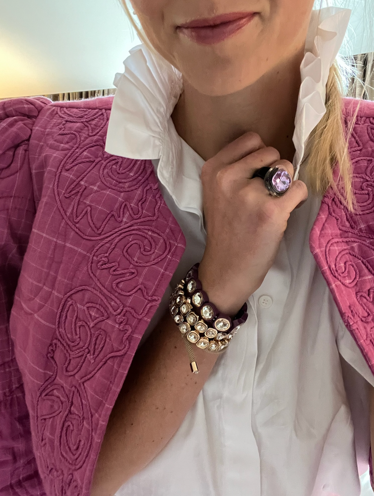 A woman wearing a white shirt and pink embroidered jacket, showcasing a large purple gemstone ring and layered crystal-embellished bracelets on their wrist.