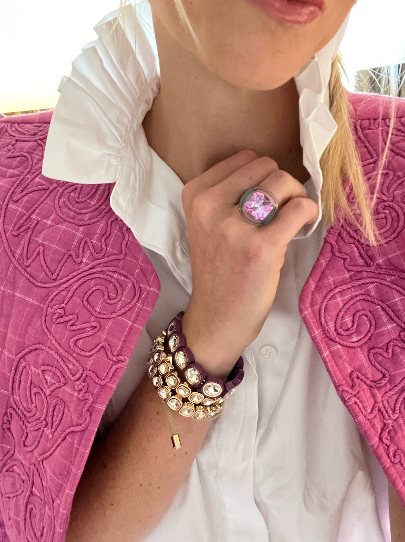 A hand adorned with stacked, purple and gold jewel-embedded bracelets and a pink gemstone ring, paired with a vibrant pink quilted vest and white blouse.