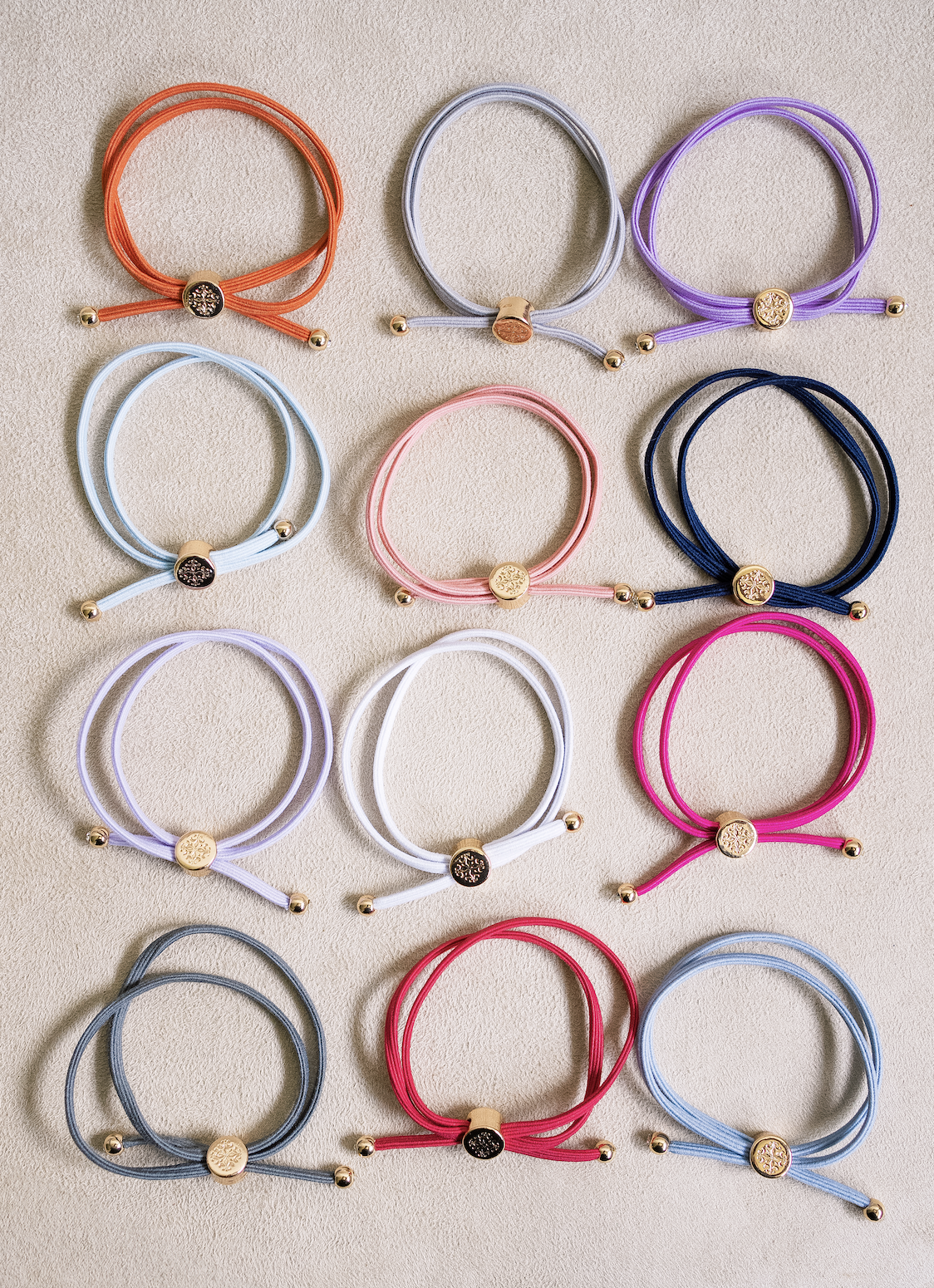 Assorted elastic hair ties with gold metal accents in vibrant colors including orange, lavender, grey, blue, pink, navy, and white, on a neutral backdrop.