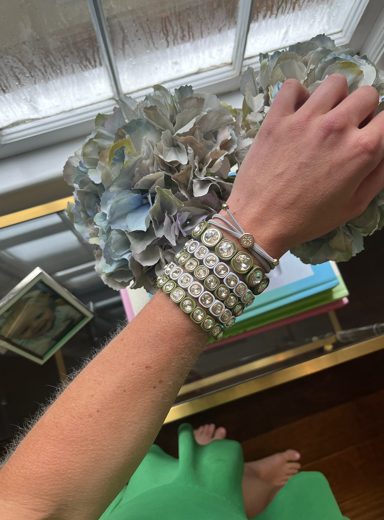 A hand adorned with multiple olive-green and beige crystal-studded stretchy bracelets and a beige hair tie, complemented by a gold-accented ring.