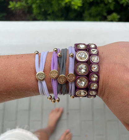 A wrist with lavender, gray, and purple double wrapped hair ties featuring gold accents, paired with two crystal-studded purple bangles.