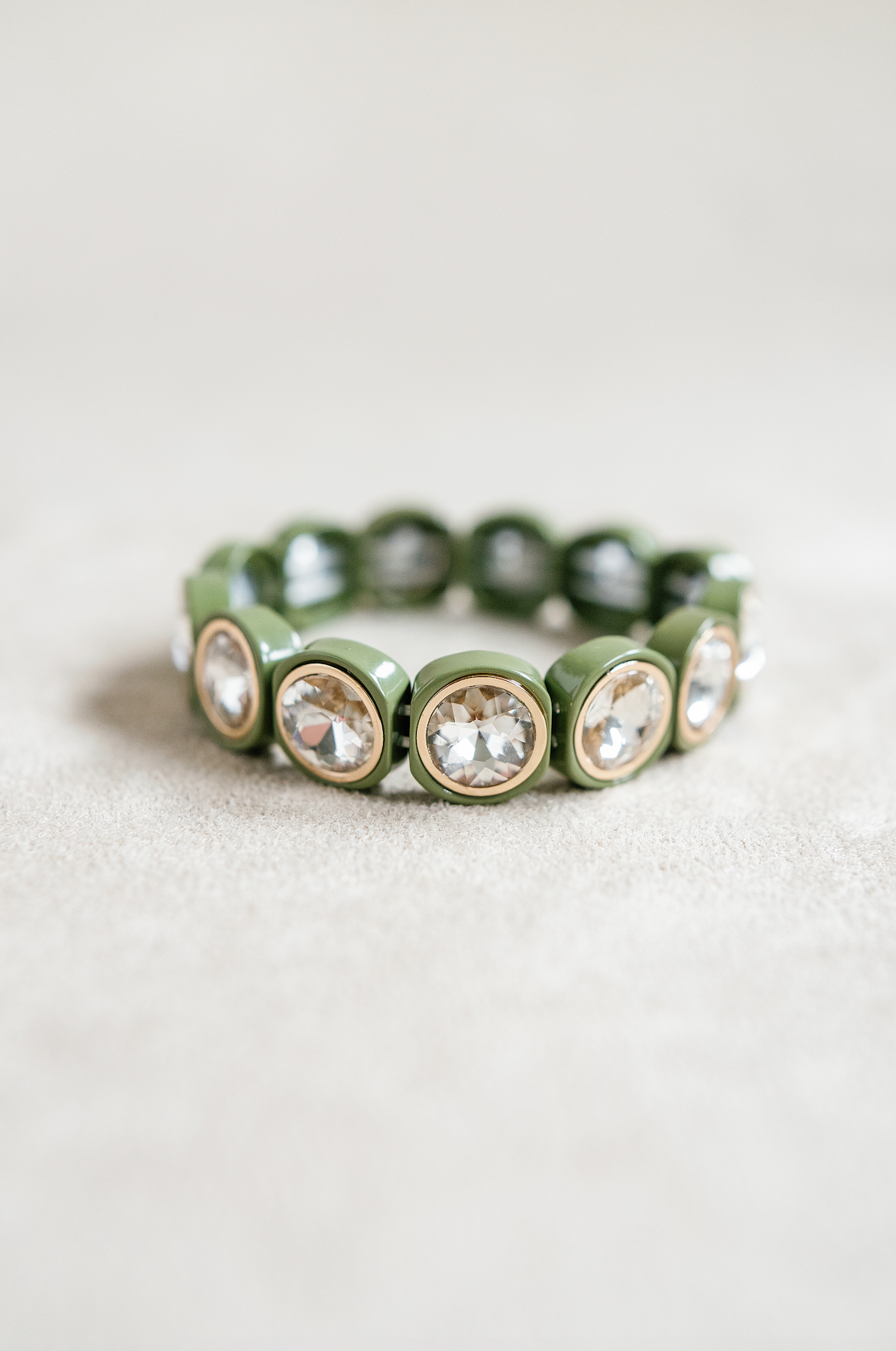 A close-up of an acrylic stretch bracelet in olive green, featuring round, crystal-like stones set in gold circular bezels that reflect light.