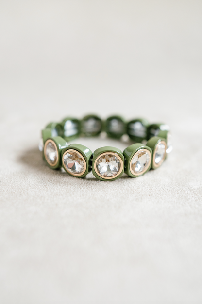 A close-up of an acrylic stretch bracelet in olive green, featuring round, crystal-like stones set in gold circular bezels that reflect light.