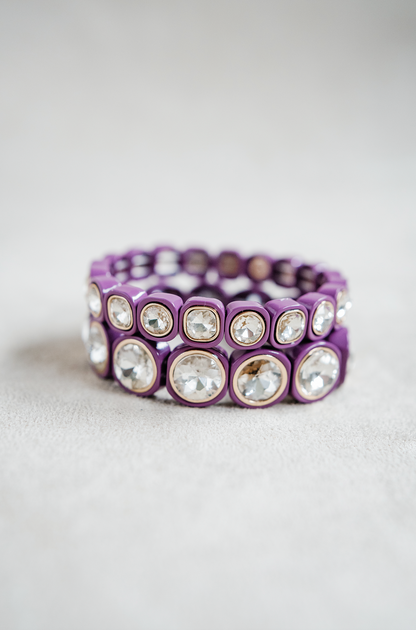 A purple bracelet features large, circular crystal stones encased in rounded gold frames, evenly spaced with elastic for a one size fits all look. Petite and round stretch bracelet stack.