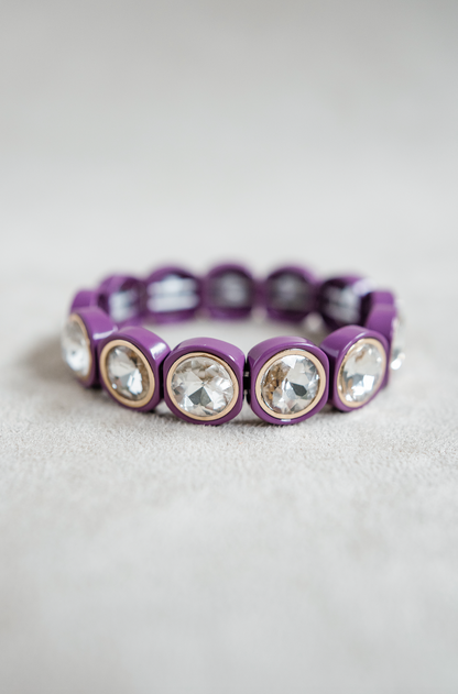 A purple bracelet features large, circular crystal stones encased in rounded gold frames, evenly spaced with elastic for a one size fits all look