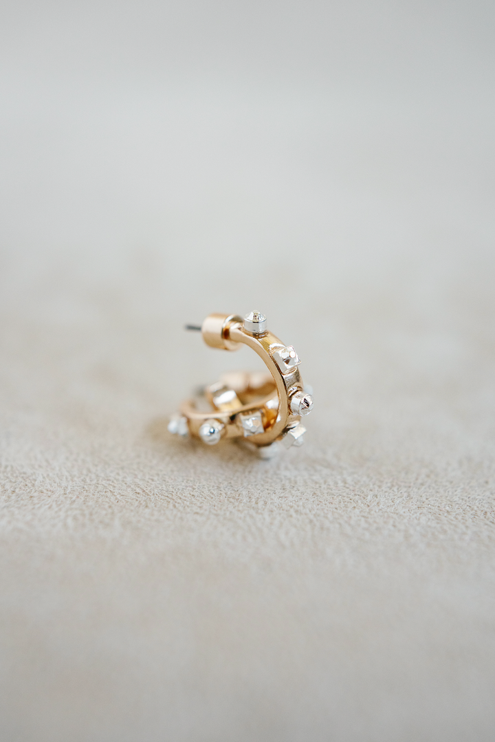 A pair of small, open hoop earrings with a gold-toned finish and silver-tipped studs surrounding the outer edge, 