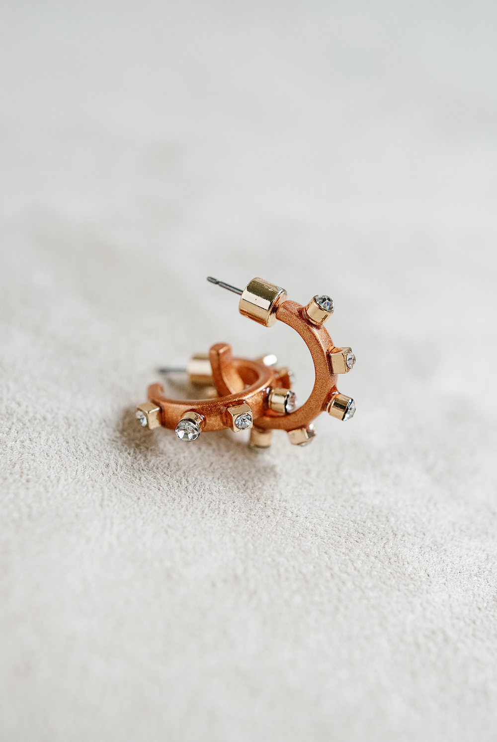 A pair of matte metal copper hoop earrings with gold accents, featuring studs around the rim and clear crystals on the post,