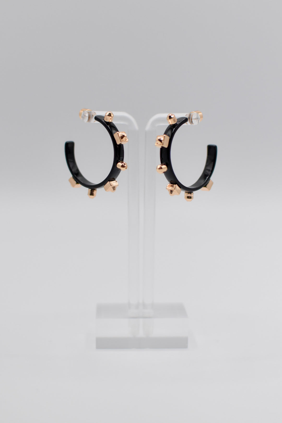 A pair of glossy, black metallic metal hoops, each adorned with six small gold studs, are mounted neatly on a transparent stand.