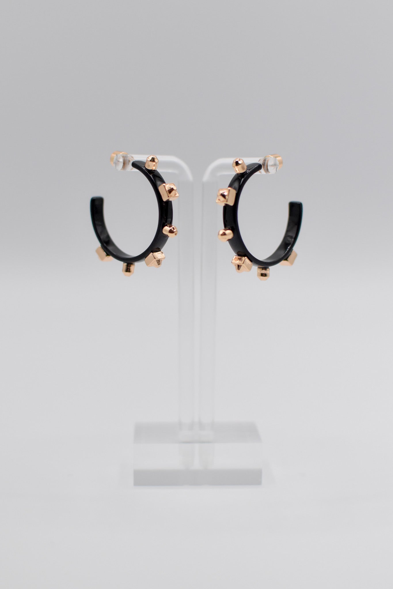 A pair of glossy, black metallic metal hoops, each adorned with six small gold studs, are mounted neatly on a transparent stand.