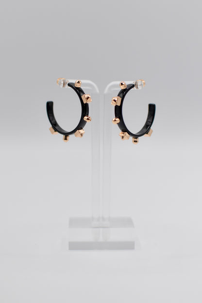 A pair of glossy, black metallic metal hoops, each adorned with six small gold studs, are mounted neatly on a transparent stand.