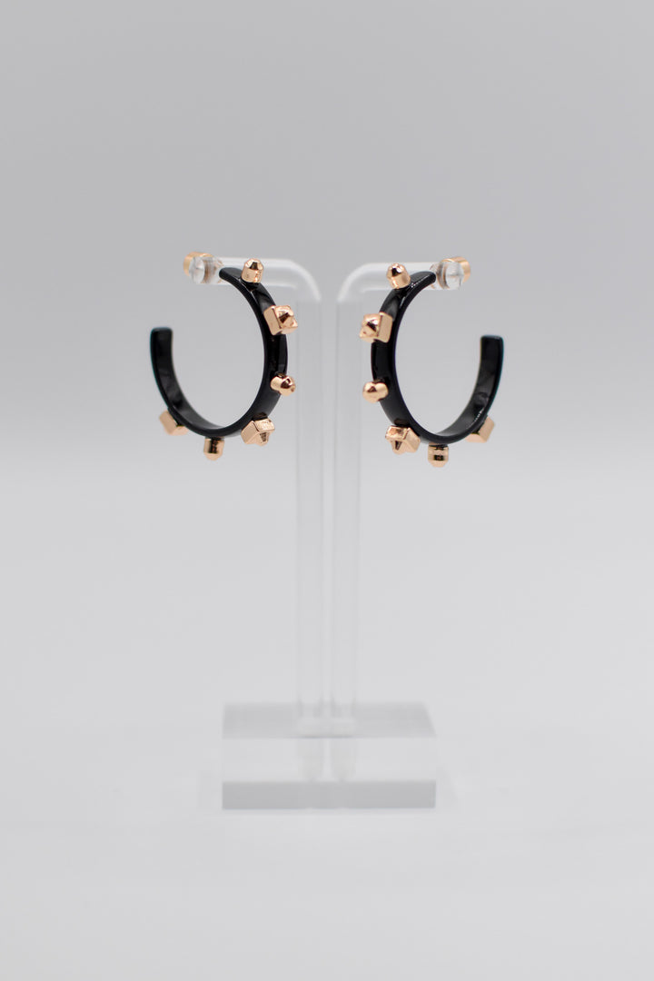 A pair of glossy, black metallic metal hoops, each adorned with six small gold studs, are mounted neatly on a transparent stand.